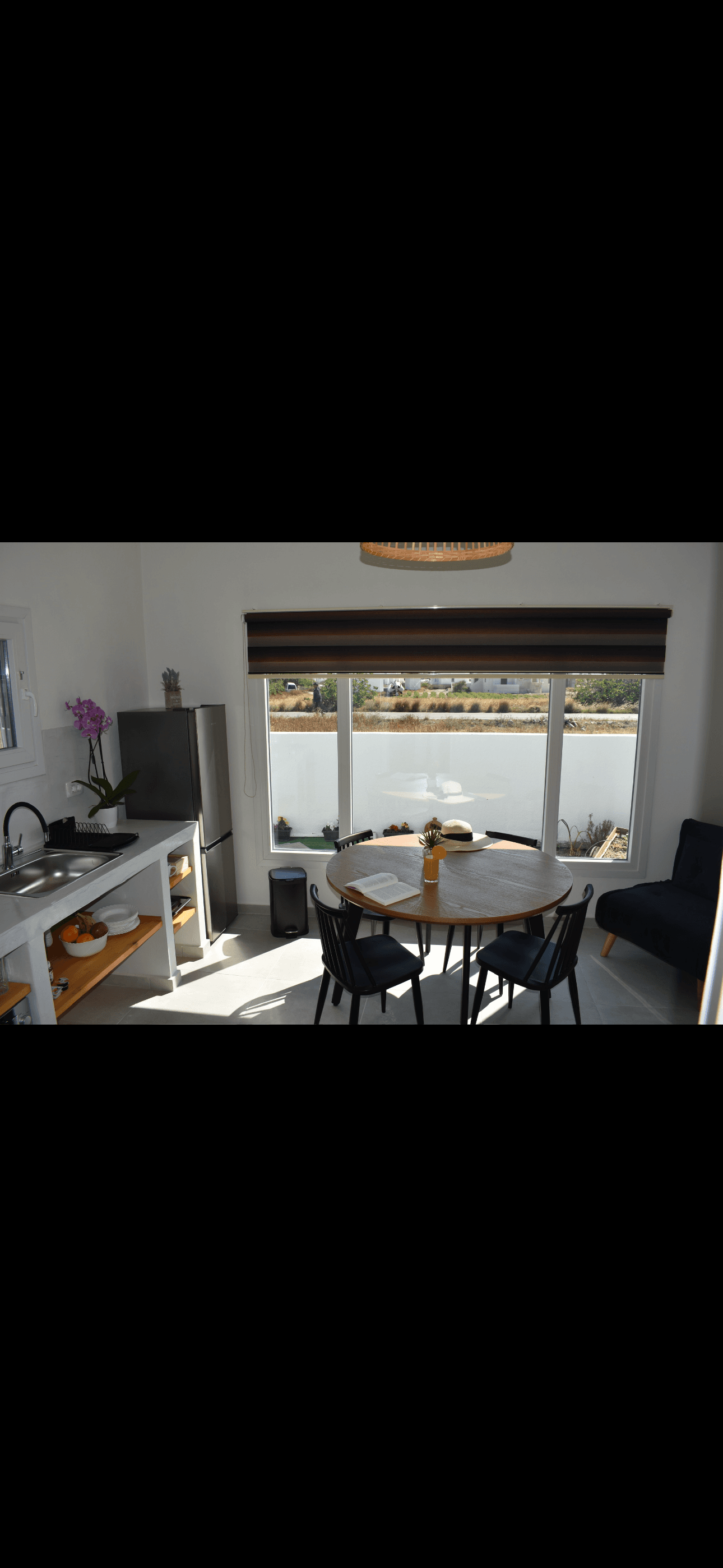 Sunlit dining area with a round wooden table, black chairs, and a large scenic window.
